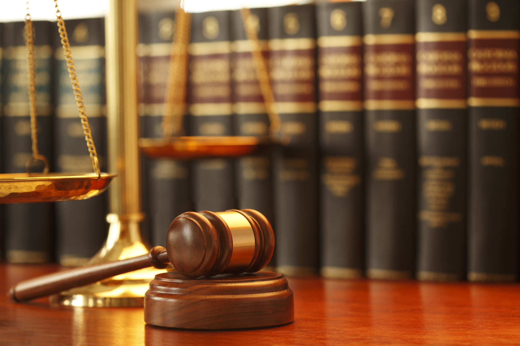 A gavel rests on sounding block next to a brass scale of justice in front of a long row of law books on a desk in a law office. Photographed with a very shallow depth of field.